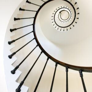 Фотография на спирално стълбище, гледано отдолу, с черни парапети и бели стени, създаваща геометричен ефект.