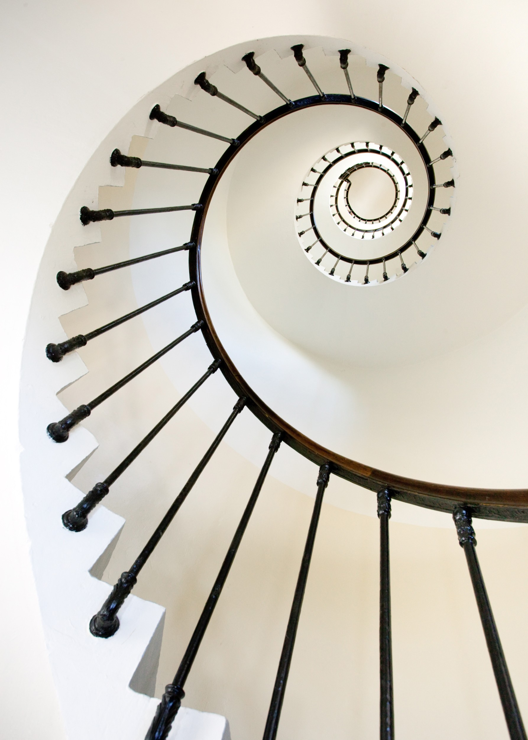 Фотография на спирално стълбище, гледано отдолу, с черни парапети и бели стени, създаваща геометричен ефект.