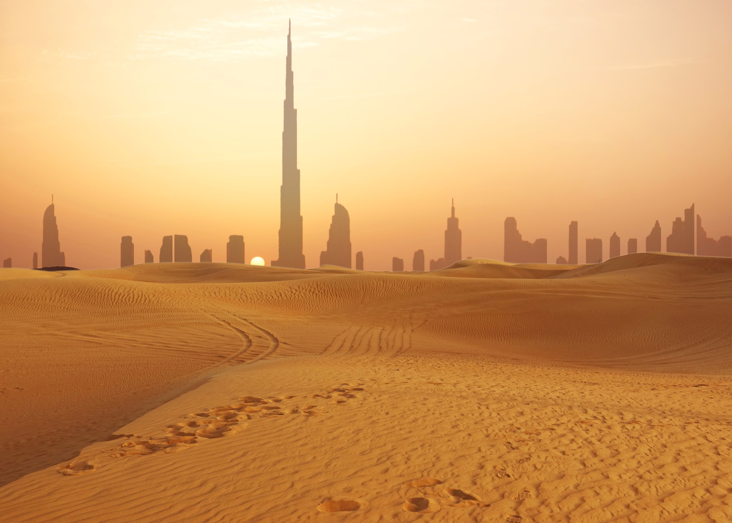 Пустиня със силует на Дубай и Бурдж Халифа при залез.