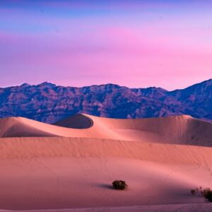 Пустинни дюни в розови и лилави цветове на залез с планини отзад.
