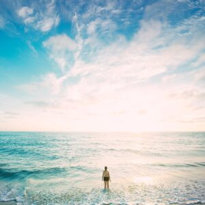 Човек стои във водата и гледа изгрева над океана